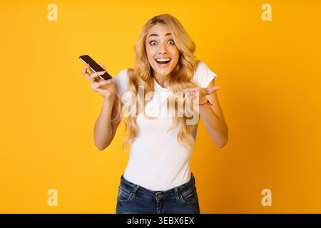 La giovane donna eccitata con i capelli lunghi mantiene il telefono su un vivace sfondo giallo Foto Stock