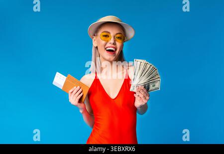 Happy Young Woman in costume da bagno in possesso di contanti e biglietto su sfondo blu Foto Stock