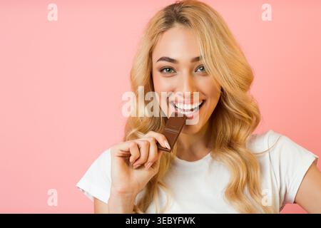 Donna sorridente che si gusta il cioccolato su uno sfondo rosa in un ambiente allegro Foto Stock