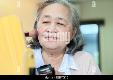 Una bella donna asiatica anziana è seduta a casa e sta facendo la sua routine mattutina di trucco. Donna anziana che si mette blush in faccia Foto Stock