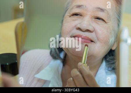 Ritratto di una bella donna anziana elegante che applica il rossetto allo specchio. Giornata nazionale del rossetto. Invecchiamento graziosamente. Messa a fuoco selettiva. Foto Stock