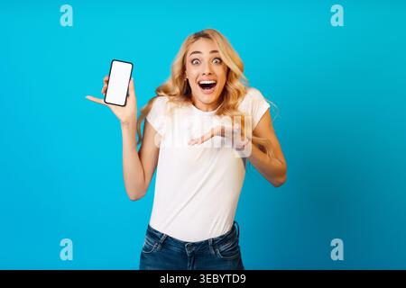 Giovane donna eccitata che tiene in mano lo smartphone su sfondo blu brillante che esprime la gioia Foto Stock