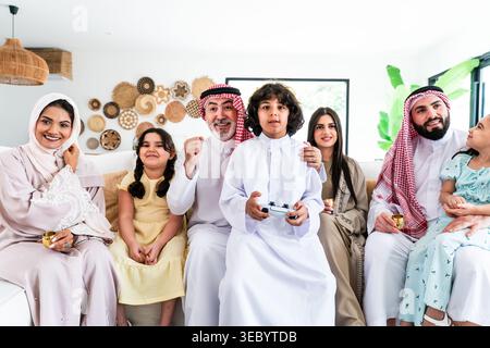 Felice famiglia araba multigenerazionale seduta su un divano nel loro moderno soggiorno, con un ragazzo che gioca ai videogiochi mentre la sua famiglia guarda e fa il tifo, Foto Stock