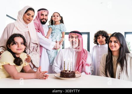 La famiglia araba si riunisce intorno a un tavolo, festeggiando un compleanno a casa, con un uomo sorridente di una torta al cioccolato con candele accese, circondato dalla sua famiglia Foto Stock