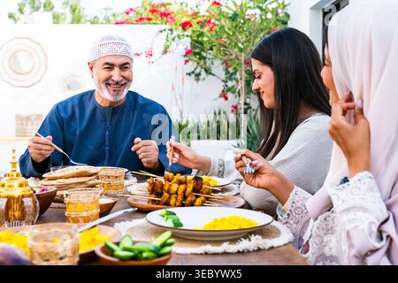 I membri della famiglia araba, tra cui padre e figlie, si riuniscono intorno a un tavolo gustando un pasto tradizionale all'aperto durante il giorno, condividendo cibo e Foto Stock