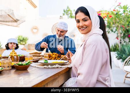 Donna araba sorridente alla macchina fotografica mentre la famiglia multi-generazionale condivide il cibo tradizionale intorno a un rustico tavolo all'aperto, godendo di insieme, ospitalità e Foto Stock