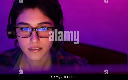 Giovane donna concentrata che indossa occhiali per giocare o svolgere attività online in una stanza poco illuminata Foto Stock
