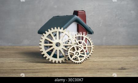Ingranaggi e ingranaggi in legno posizionati di fronte a una casa di giocattoli in miniatura che rappresenta i concetti di automazione domestica, ingegneria e costruzione residenziale. Foto Stock