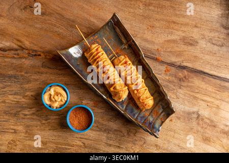 Una vista dall'alto verso il basso di due cani di mais perfettamente dorati, splendidamente conditi con salsa e conditi. Accompagnato da piccole ciotole di condime saporito Foto Stock