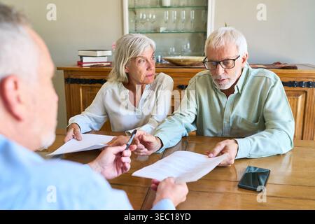 Una coppia senior si impegna in una discussione mirata con un consulente finanziario, esaminando documenti importanti per la pianificazione pensionistica e la gestione immobiliare. Foto Stock