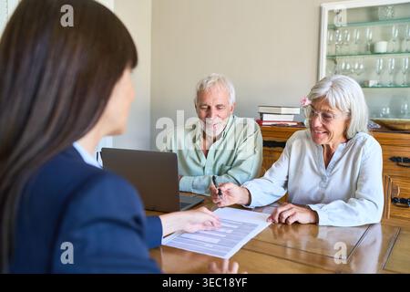 Una coppia di anziani si occupa di pianificazione pensionistica e protezione finanziaria, firmando documenti importanti con un consulente professionale a un tavolo nella loro h Foto Stock