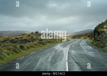 Strada di montagna curvilinea N71 che attraversa il suggestivo paesaggio glaciale della contea di Kerry, in Irlanda, in una giornata nebbiosa. Foto Stock