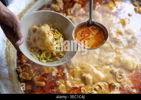 Persona che tiene la ciotola della tradizionale zuppa di spaghetti al curry per preparare il pasto, versando un ricco brodo d'arancia sul pollo da un grande vaso bollente con mestolo da portata Foto Stock