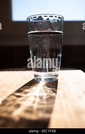 Un bicchiere sfaccettato di acqua pura su una superficie di legno chiaro. La luce naturale del sole crea splendidi motivi caustici e ombre spettacolari. Perfetto per la salute, W Foto Stock