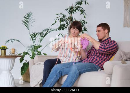Durante una visita casalinga, un figlio adulto apprende il mestiere della maglieria da sua madre, sfidando gli stereotipi di genere sul loro comodo divano. Un ricco Foto Stock