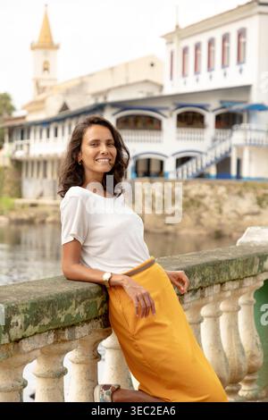 Una donna si trova in riva al fiume, appoggiata a una ringhiera in pietra in un affascinante villaggio. Indossa un top bianco e una gonna gialla, con bui pittoreschi Foto Stock