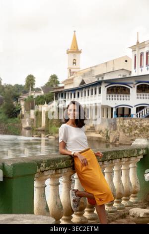 Una donna si trova in riva al fiume, appoggiata a una ringhiera in pietra in un affascinante villaggio. Indossa un top bianco e una gonna gialla, con bui pittoreschi Foto Stock