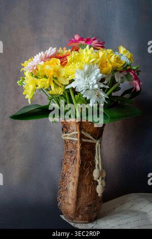 Vivace bouquet di fiori gialli, bianchi e rosa in un vaso di argilla marrone testurizzato, adornato con spago e perle su sfondo scuro. Foto Stock