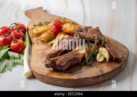 Costolette di agnello grigliate servite con patate arrosto, aglio e pomodori ciliegini freschi su un rustico bordo di legno. Foto Stock