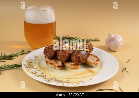 Salsicce rumene mici senza pelle alla griglia condite con senape, servite su fette di pane tostato accanto a un bicchiere di birra fredda e a un bulbo d'aglio. Foto Stock