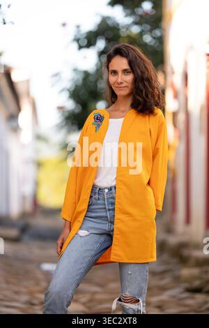 Una donna si trova in una strada acciottolata in una vivace città. Indossa un cappotto giallo su una camicia bianca e jeans blu. Viene visualizzato lo sfondo Foto Stock