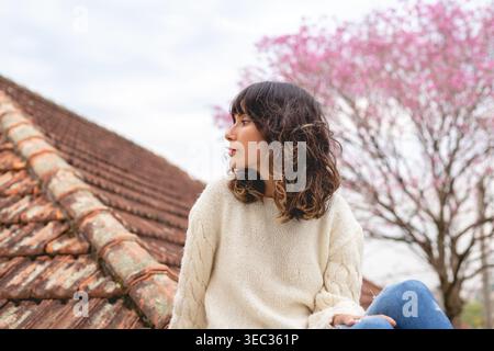 Una giovane donna gode della vista mentre siede sul tetto. Ha i capelli ricci e indossa un maglione leggero. Un albero in fiore di ciliegio aggiunge colore al dorso Foto Stock