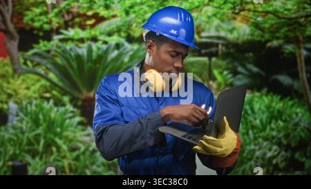 Uomo operaio edile in elmetto blu che tiene in mano un computer portatile e una chiave, che stringe la mano con i guanti nella foresta; frustrazione nella risoluzione dei problemi. Foto Stock