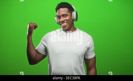 L'uomo nero che indossa le cuffie si flette bicipite destro e sorride verso la fotocamera in uno studio green screen indossando una t shirt bianca; un ritmo sicuro. Foto Stock