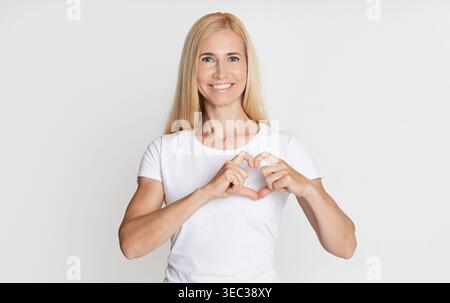 Donna sorridente con lunghi capelli biondi che creano una forma a cuore con le mani in casa Foto Stock