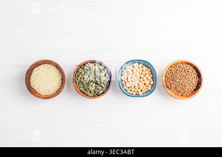 Cereali assortiti, semi e legumi in ciotole di legno disposte su sfondo bianco, concetto di cibo sano. Vista dall'alto, disposizione piatta. Foto Stock