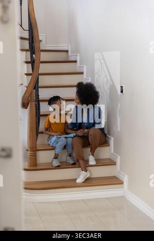 Madre e bambino seduti su scale curve di legno a casa, leggendo un quaderno aperto Foto Stock