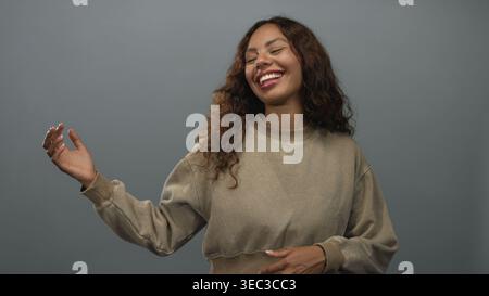 Donna sorridente con gesti della mano e posa rilassata in uno studio grigio con felpa beige e ondeggiante delicatamente; gioia spensierata. Foto Stock