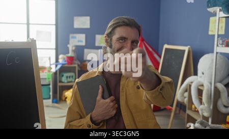 Uomo che indossa una giacca gialla che tiene il tablet sorride e punta le dita verso la fotocamera in classe; incoraggia la fiducia della connessione. Foto Stock