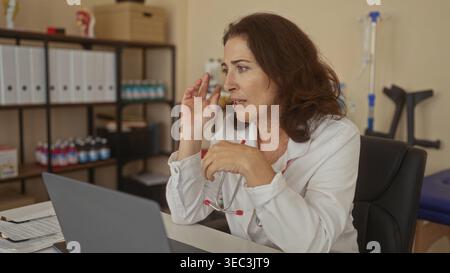 Donna medico ispanico di mezza età che indossa un cappotto bianco con stetoscopio, gesti con le mani mentre si consulta al computer portatile nell'edificio; compassione. Foto Stock