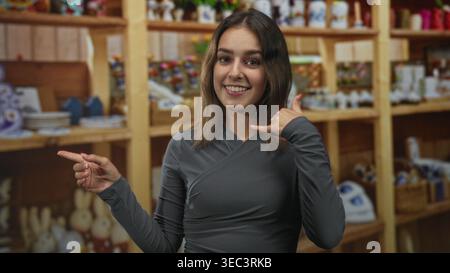 Donna che punta il dito sugli scaffali e mano a mano per un gesto di chiamata in negozio, sorridendo con la collana visibile; invito cordiale. Foto Stock