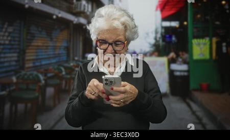 Donna anziana nonna dai capelli bianchi che indossa gli occhiali che reggono lo smartphone, tocca lo schermo con i pollici in un bar di strada con sedie e graffiti; digita Foto Stock