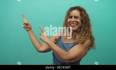 Una donna di mezza età punta entrambe le dita indice verso l'alto in studio, sorridendo ampiamente con denti visibili e capelli ricci; gioiosa fiducia. Foto Stock