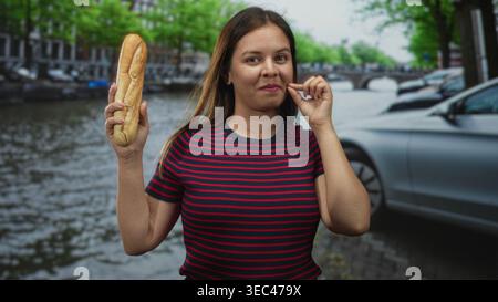Donna che tiene la baguette e le onde mano sulla strada del canale di amsterdam mentre sorride in grande; viaggio divertente. Foto Stock