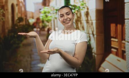 Donna che presenta le palme sorridente come se tenesse un oggetto in una stretta e soleggiata strada acciottolata incorniciata da muri in pietra e porta in legno; benvenuto Foto Stock