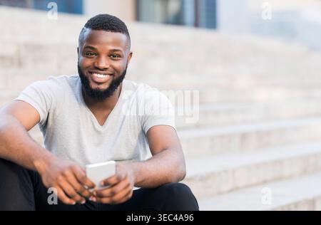 Giovane sorridente mentre si siede sui gradini all'aperto e utilizza lo smartphone durante il giorno Foto Stock