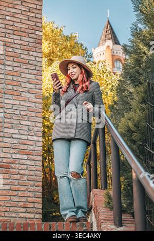 Giovane donna con un cappello e un blazer in piedi su scale di mattoni, con un telefono cellulare, circondata dal fogliame autunnale in un parco cittadino di Belgrado Foto Stock