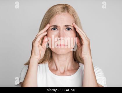 Donna con capelli biondi visibilmente stressata mentre tiene la testa in un ambiente neutro Foto Stock
