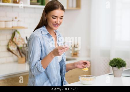 Donna che si diverte a fare colazione mentre usa lo smartphone in un ambiente moderno da cucina Foto Stock