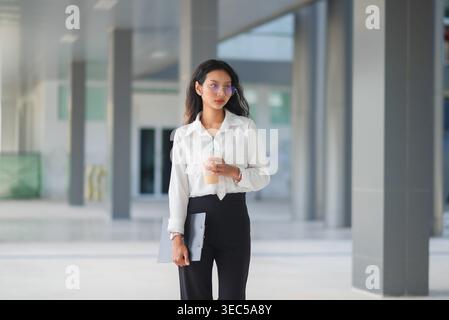 Ritratto a figura intera di una donna professionista che tiene in mano caffè ghiacciato e appunti all'esterno di un edificio moderno, che trasmette fiducia, mobilità sul posto di lavoro, AN Foto Stock