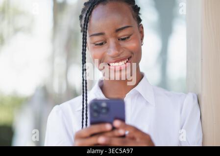 Giovane donna d'affari nera sorridente allo smartphone mentre si appoggia all'aperto vicino a un edificio moderno. Messaggistica online, navigazione mobile, comunicazioni digitali Foto Stock