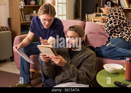 Giovani studenti adulti che studiano insieme in uno spazio abitativo condiviso, leggono appunti e scrivono compiti. Utile per marketing didattico, vita universitaria, lavoro di squadra, preparazione agli esami Foto Stock