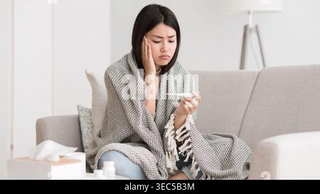 Donna seduta sul divano con coperta che controlla la temperatura durante la malattia a casa in inverno Foto Stock