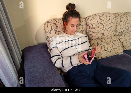 Donna che si rilassa sul divano di casa, ascolta musica con le cuffie e tiene in mano un lettore di cassette analogico personale Foto Stock