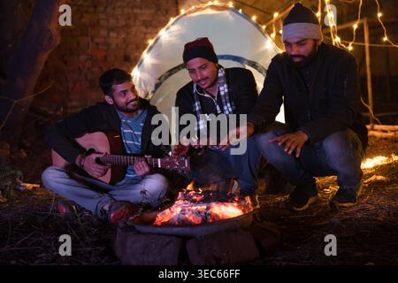 Un gruppo di tre amici condivide una serata memorabile intorno a un falò, ascoltando la musica di una chitarra e battendo ritmici sotto il cielo stellato. Foto Stock