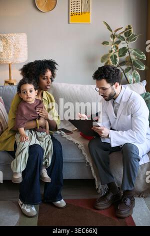 Shot verticale di madre nera che tiene in braccio il bambino mentre un medico di sesso maschile scrive appunti durante la visita a casa Foto Stock